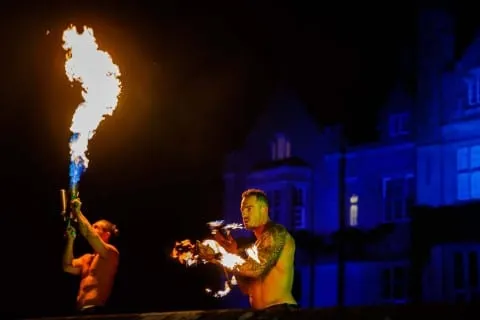 Fire performers using a flame thrower and fire staff outdoors at dusk